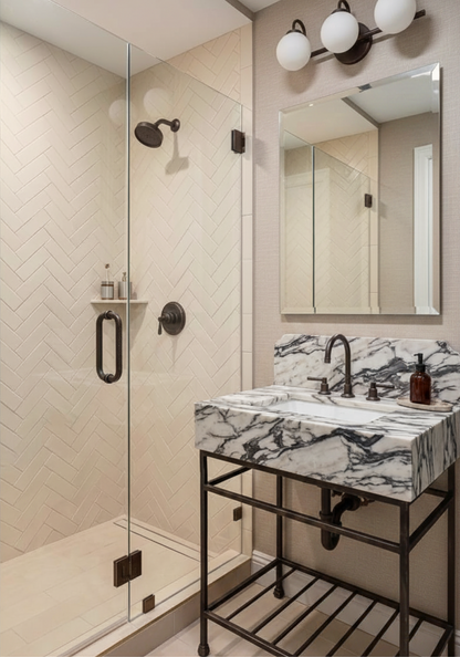 A modern bathroom vanity setup featuring a black metal frame sink with a white and gray marble countertop, a matching marble-look basin, and a large wall mirror. To the left is a walk-in shower with beige herringbone tile walls and dark bronze fixtures.
