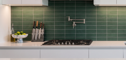 A modern kitchen scene with glossy green subway tiles on the backsplash, white cabinetry, a black gas cooktop, a stainless steel faucet, and countertop accessories including a knife block, bowl, and bottle.