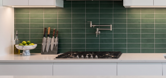 A modern kitchen scene with glossy green subway tiles on the backsplash, white cabinetry, a black gas cooktop, a stainless steel faucet, and countertop accessories including a knife block, bowl, and bottle.