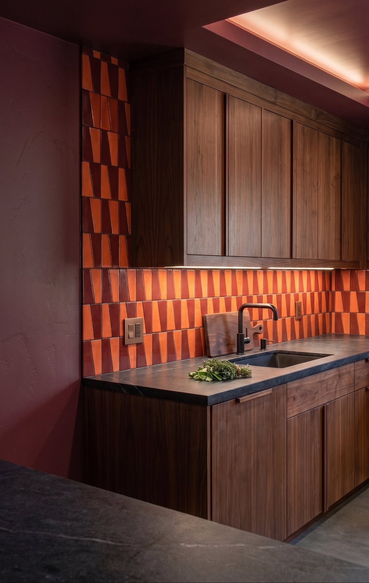 A modern kitchen corner with dark wood cabinets, a dark countertop, and a bold orange and dark brown geometric patterned tile backsplash.