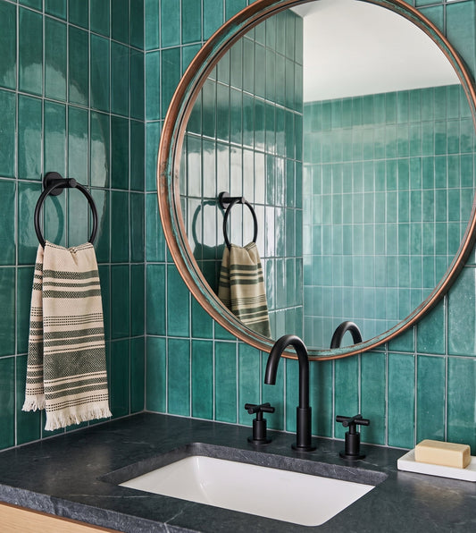 A modern bathroom features a round copper-rimmed mirror, vibrant green zellige tile walls, and a black faucet paired with a striped hand towel on a ring.