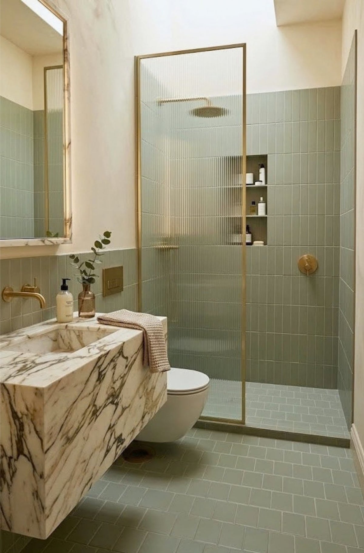 A modern bathroom features sage green tiles, a marble vanity, and a fluted glass walk-in shower with brass fixtures.