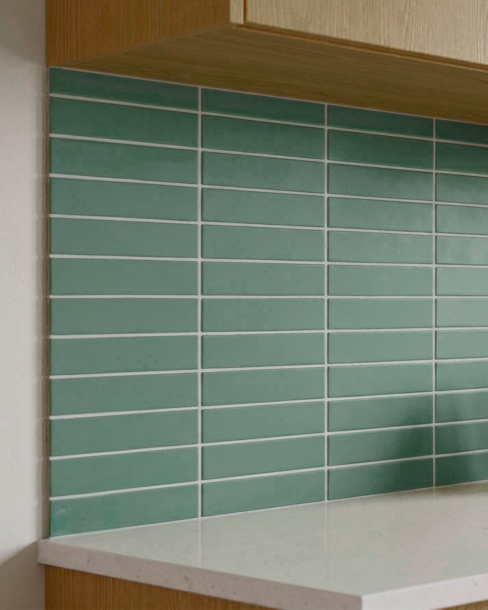 Green rectangular subway tiles with white grout lines arranged on a kitchen backsplash behind a white countertop and light wood cabinets.