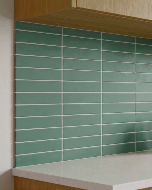 Green rectangular subway tiles with white grout lines arranged on a kitchen backsplash behind a white countertop and light wood cabinets.