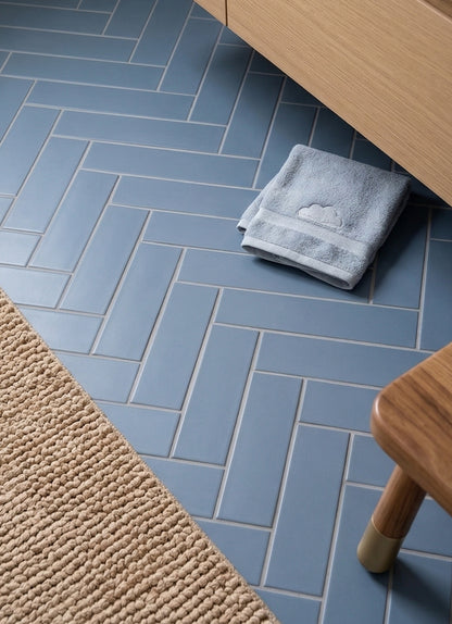 Denim blue 2x8 clay tiles by Clay Imports in a herringbone pattern on a floor, with a blue towel and wooden stool.