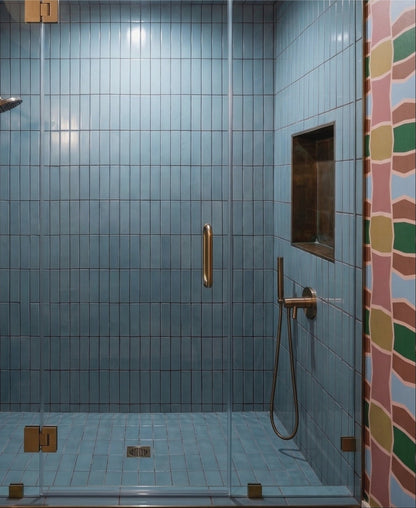 A modern walk-in shower enclosure with light blue square tiles, a clear glass door, and brass hardware, shown in a real bathroom setting with a patterned tile strip on the right.