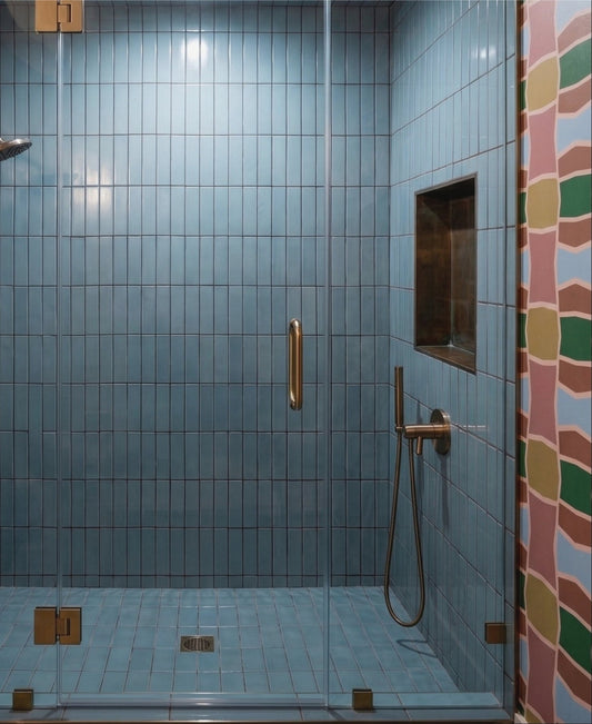 A modern walk-in shower enclosure with light blue square tiles, a clear glass door, and brass hardware, shown in a real bathroom setting with a patterned tile strip on the right.