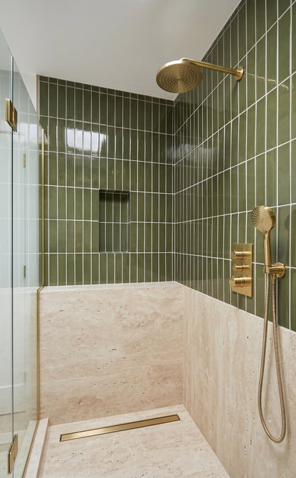 A modern walk-in shower stall with glossy green vertical wall tiles, a beige stone floor and bench, and brushed brass fixtures including a rain showerhead, handheld shower, and control valves.