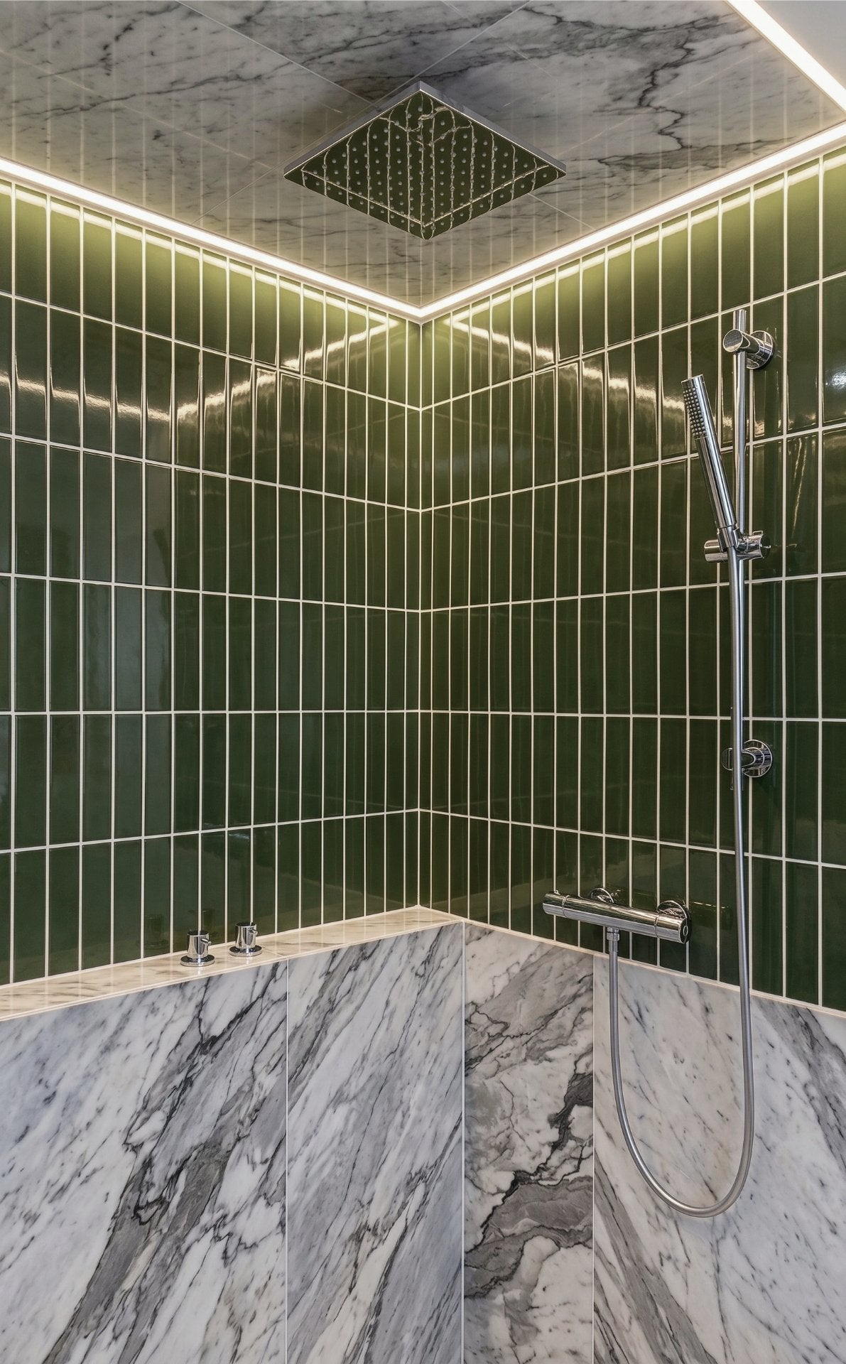 A modern bathroom corner with glossy dark green wall tiles, a marble-look bathtub or bench, a chrome handheld shower, and warm LED strip lighting along the top edge.