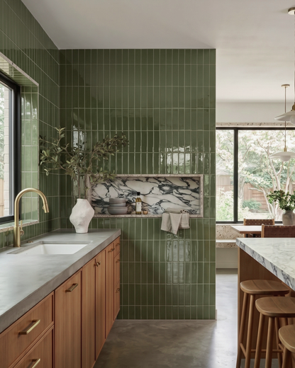 A modern kitchen features glossy olive green vertical subway tiles, wooden cabinets with a concrete countertop, and a marble-backed niche displaying decorative bowls.