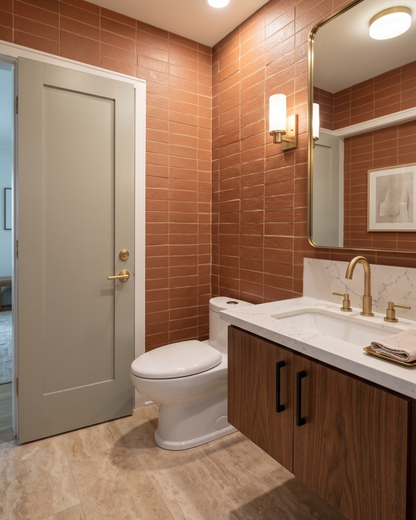 A modern bathroom vanity featuring a white marble countertop with a gold faucet, a white toilet, and a door, set against warm terracotta tiled walls and light wood cabinets.