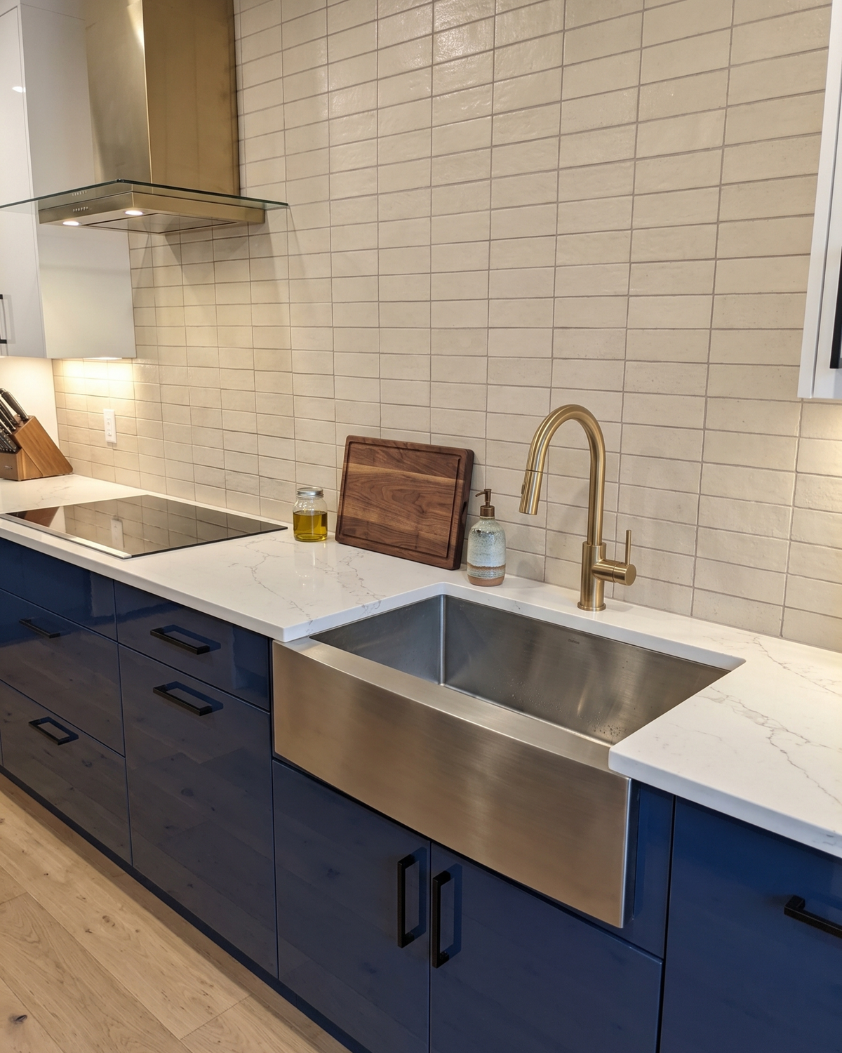 A modern kitchen setup featuring a large stainless steel farmhouse sink with a high-arc brass faucet, set into white marble countertops and navy blue cabinets.