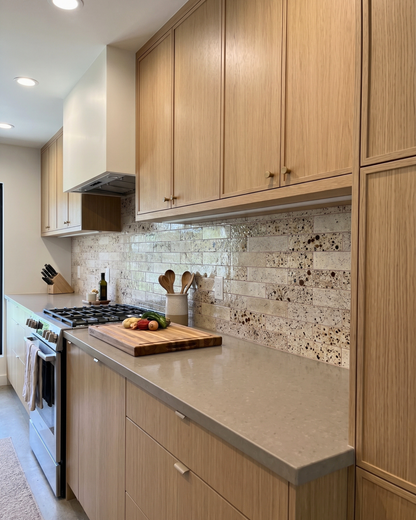 A modern kitchen interior with light wood cabinets, a speckled stone backsplash, gray countertops, a built-in gas stove, and a wooden cutting board with fruit on the counter.