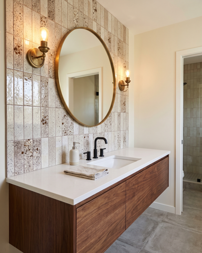 A modern bathroom vanity featuring a floating wood cabinet with a white countertop, integrated sink, black faucet, and a large oval mirror. The wall behind the vanity is covered in textured beige and brown mosaic tiles, illuminated by two brass sconces.