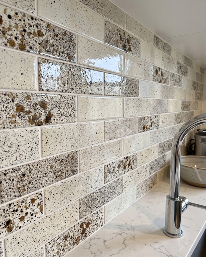 Glossy rectangular ceramic subway tiles with a speckled terrazzo pattern in cream, beige, and brown, installed on a kitchen backsplash above a white marble countertop with a chrome faucet.
