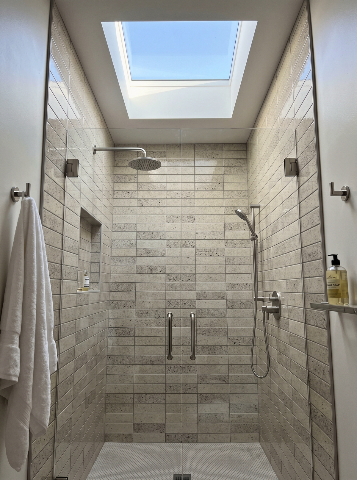 A modern walk-in shower enclosure with light beige stone-look rectangular tiles, a glass door, chrome fixtures, a built-in niche, a hanging white towel, and a skylight above.