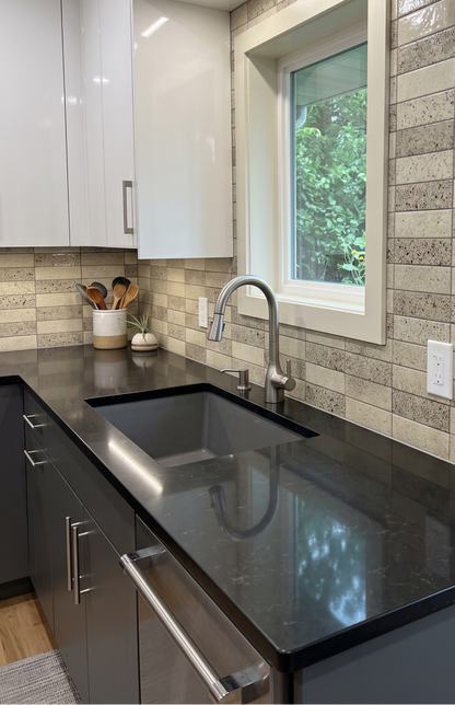 A modern kitchen sink area with a dark charcoal countertop, a stainless steel gooseneck faucet, a black undermount sink, and a beige stone tile backsplash. White upper cabinets and a window looking out onto green trees are visible.