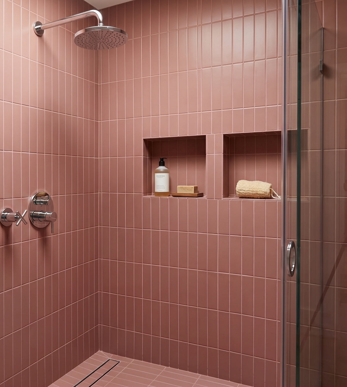 A modern shower stall with pink rectangular wall tiles, a glass door, built-in shelves with toiletries, and a round showerhead.