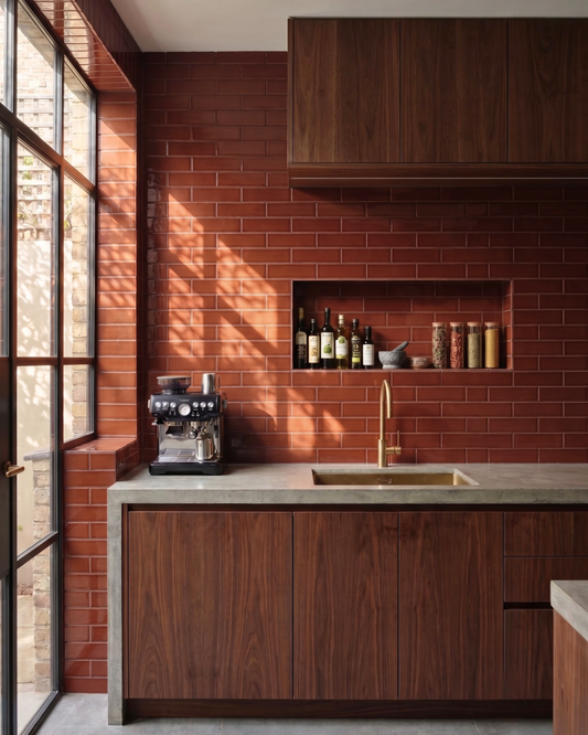 A modern kitchen interior with dark wood cabinets, a red brick backsplash, a concrete countertop, and a brass sink faucet. A built-in shelf holds bottles and jars, and a window on the left casts warm sunlight across the wall.