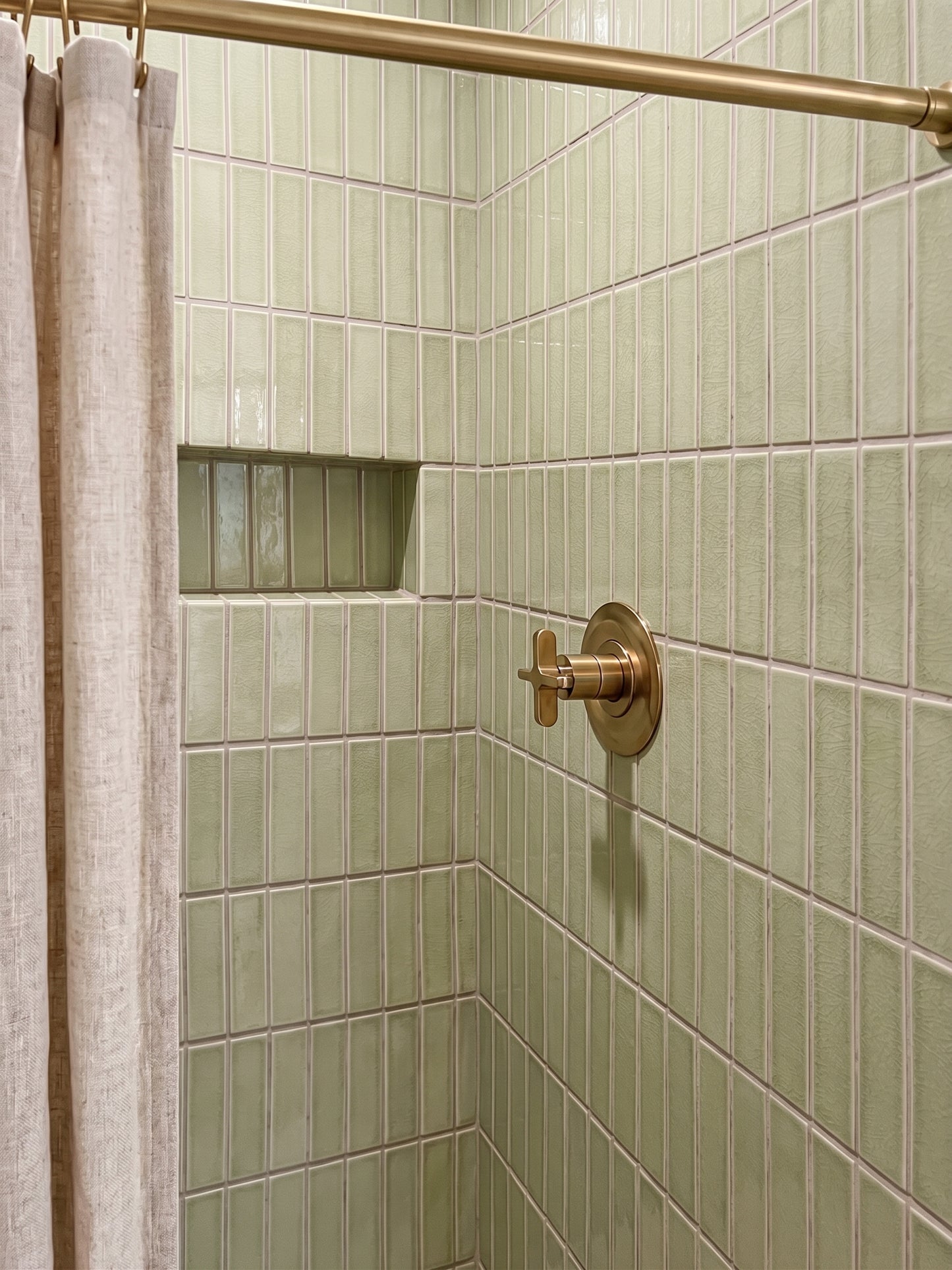 A close-up of a tiled shower corner with pale green rectangular wall tiles, a brass shower rod and control trim, and part of a beige shower curtain visible on the left.