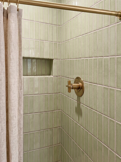 A close-up of a tiled shower corner with pale green rectangular wall tiles, a brass shower rod and control trim, and part of a beige shower curtain visible on the left.