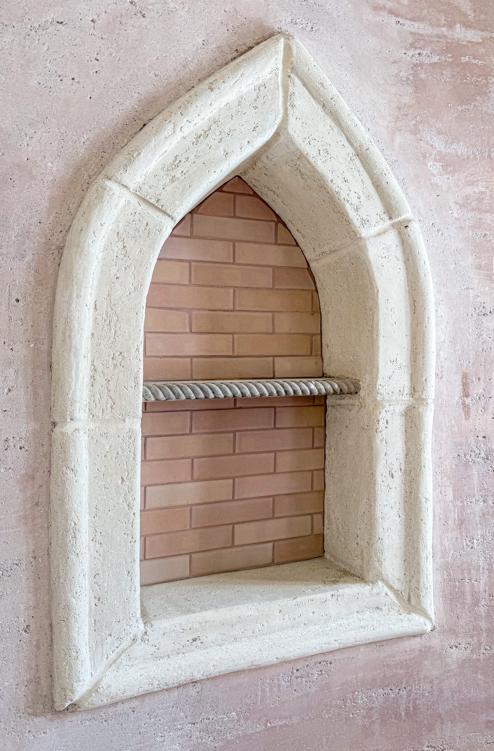 An arched stone niche with a textured off-white frame, featuring a built-in brick shelf and a rope rack, set against a textured pinkish-beige plaster wall.