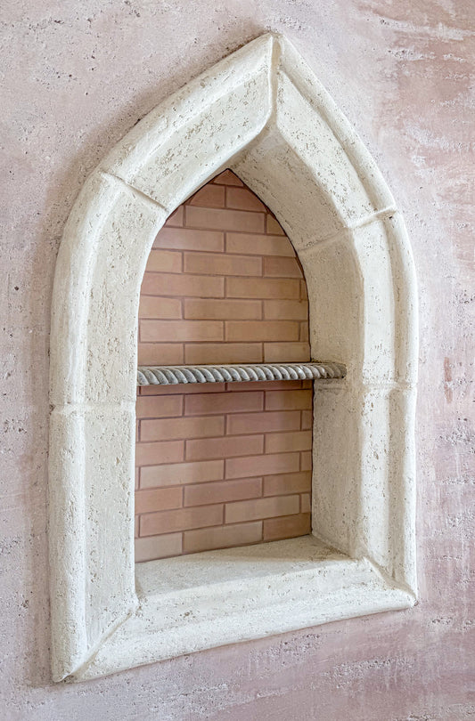 An arched stone niche with a textured off-white frame, featuring a built-in brick shelf and a rope rack, set against a textured pinkish-beige plaster wall.