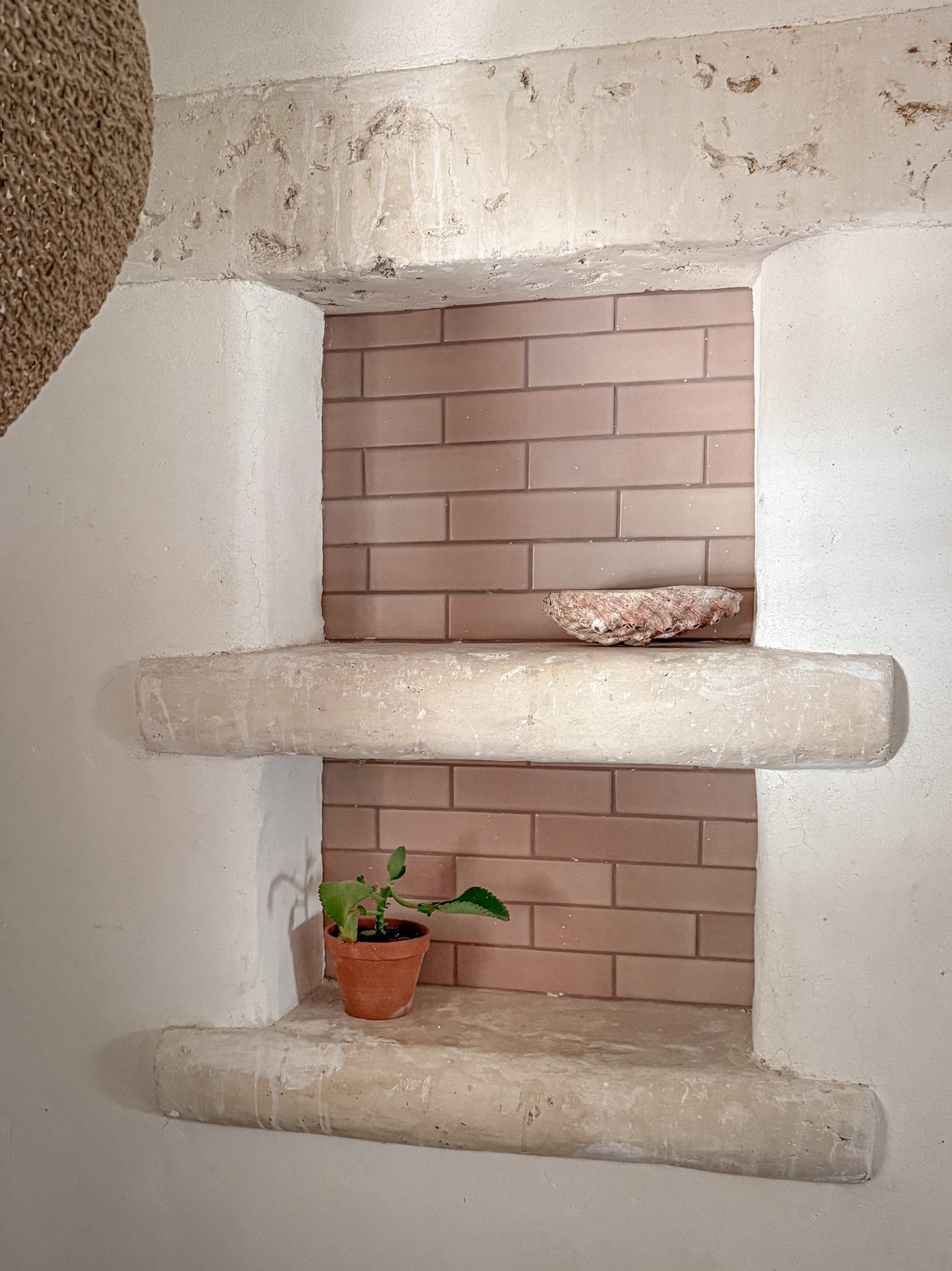 A rustic two-tiered stone shelf niche set into a white plaster wall, featuring a pinkish-beige brick backsplash and styled with a small potted plant and a decorative bowl.