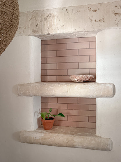 A rustic two-tiered stone shelf niche set into a white plaster wall, featuring a pinkish-beige brick backsplash and styled with a small potted plant and a decorative bowl.