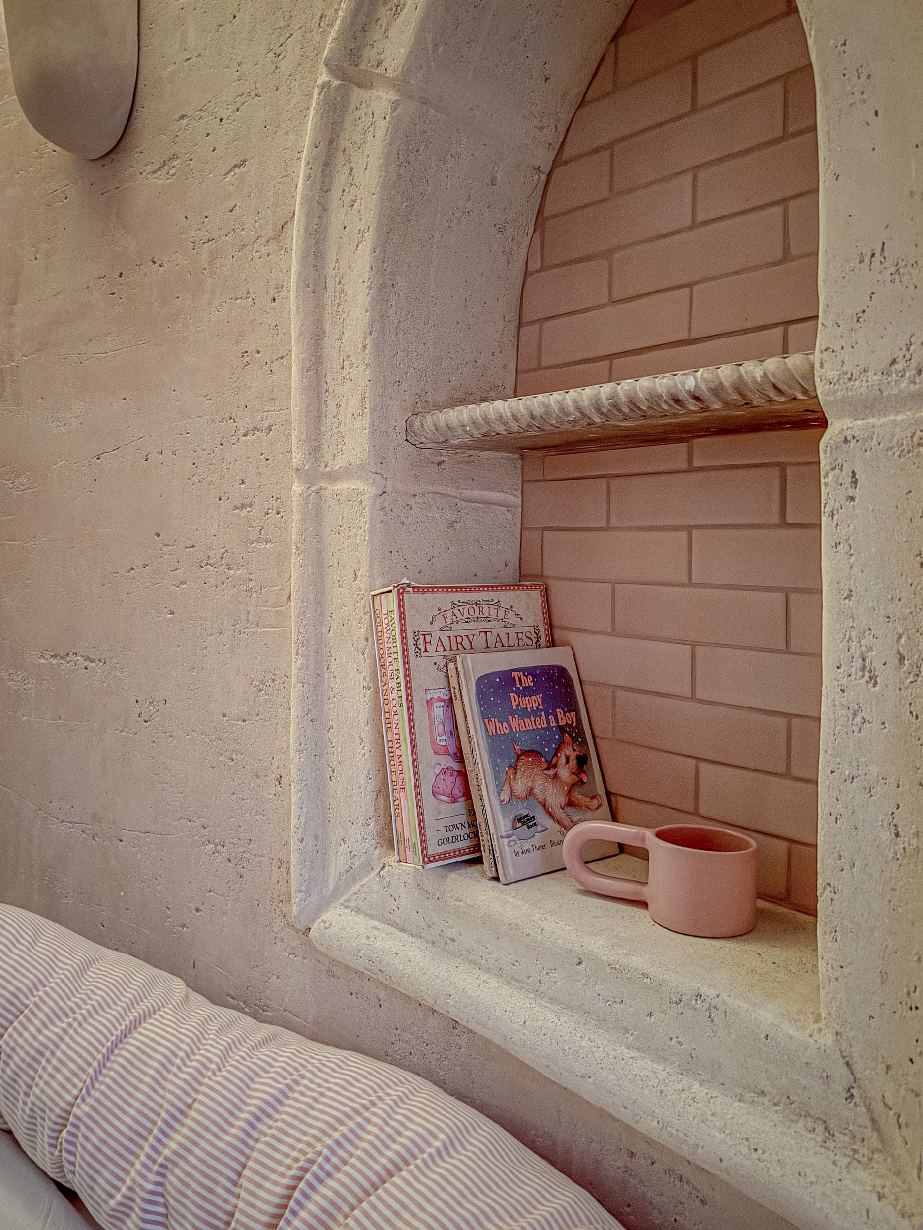 A cozy interior scene showing a built-in stone niche with a pink brick backing, a rope shelf, a stack of children's books, and a pink mug resting on the ledge.