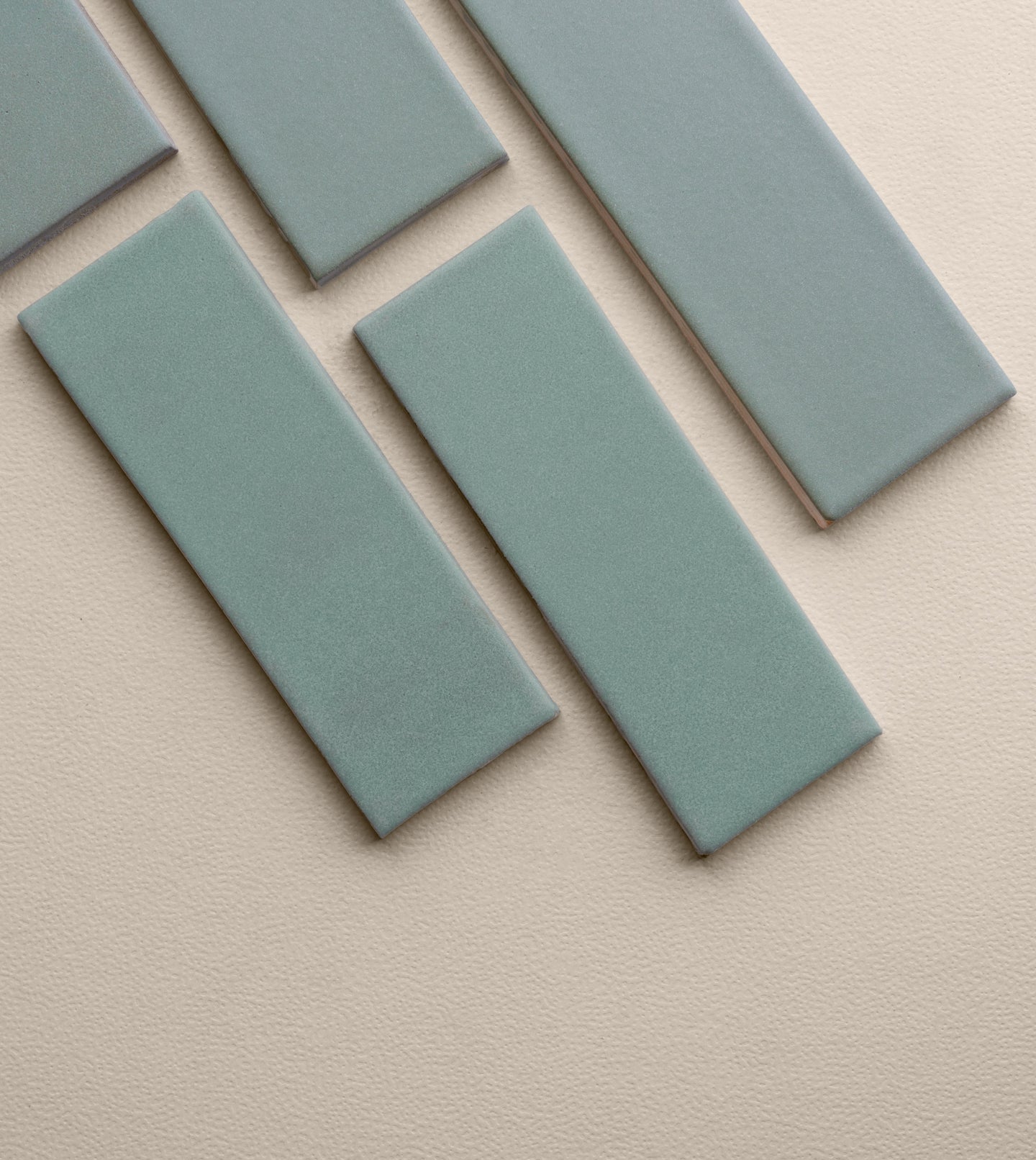 Muted blue-green 2x6 clay tile by Clay Imports, with a matte, textured surface, showing light brown edges, arranged diagonally on a light beige background.