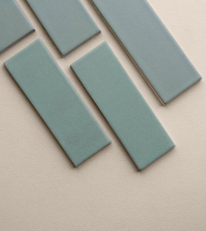 Muted blue-green 2x6 clay tile by Clay Imports, with a matte, textured surface, showing light brown edges, arranged diagonally on a light beige background.