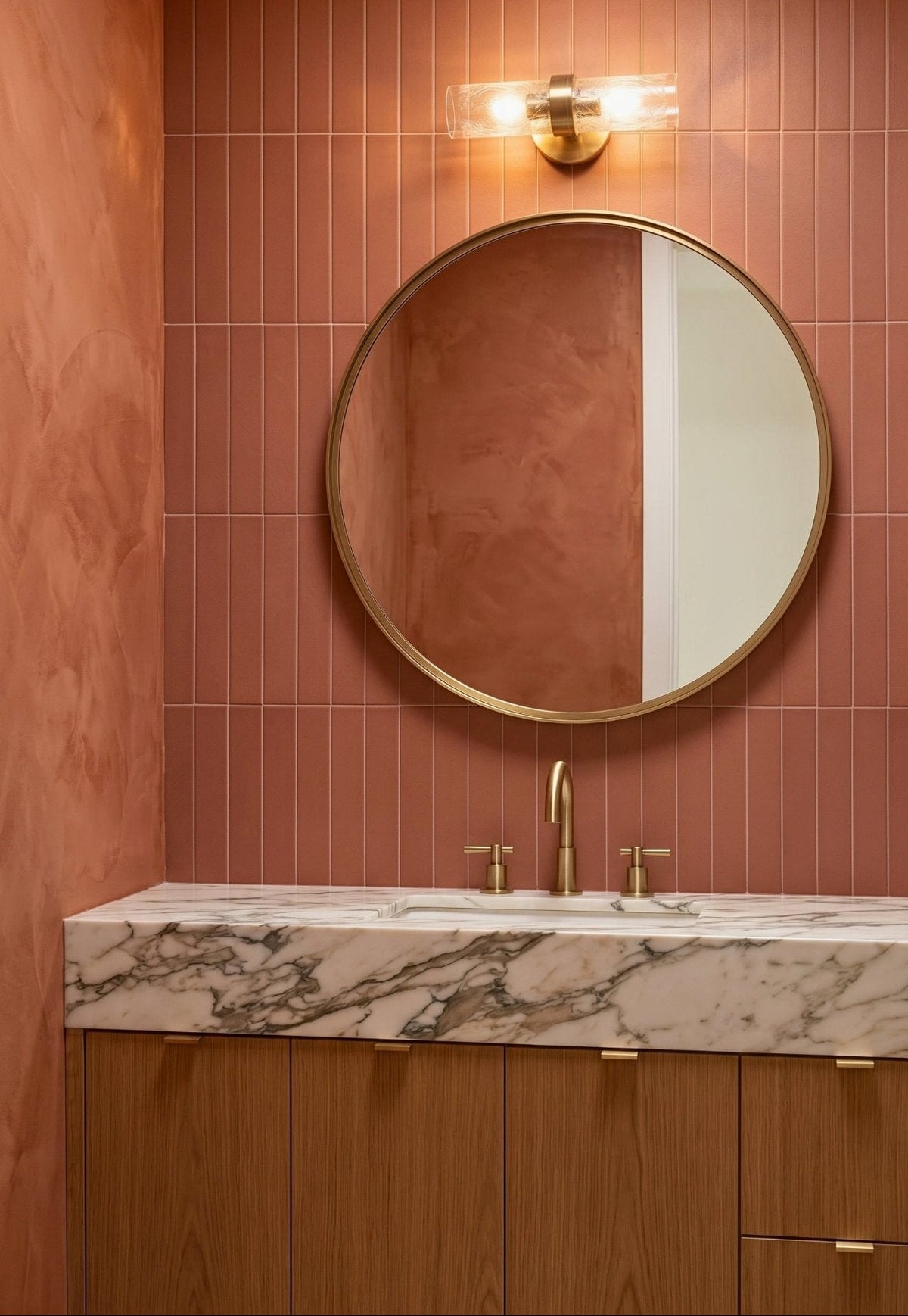A round brass-framed mirror hangs above a marble-topped vanity with brass fixtures against a terracotta tile wall in a modern bathroom.