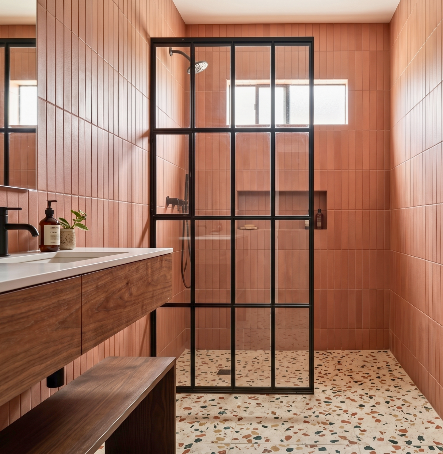 A modern bathroom interior with warm terracotta tiled walls, a wood vanity with a white sink, a black-framed shower glass enclosure, and a terrazzo-style floor.