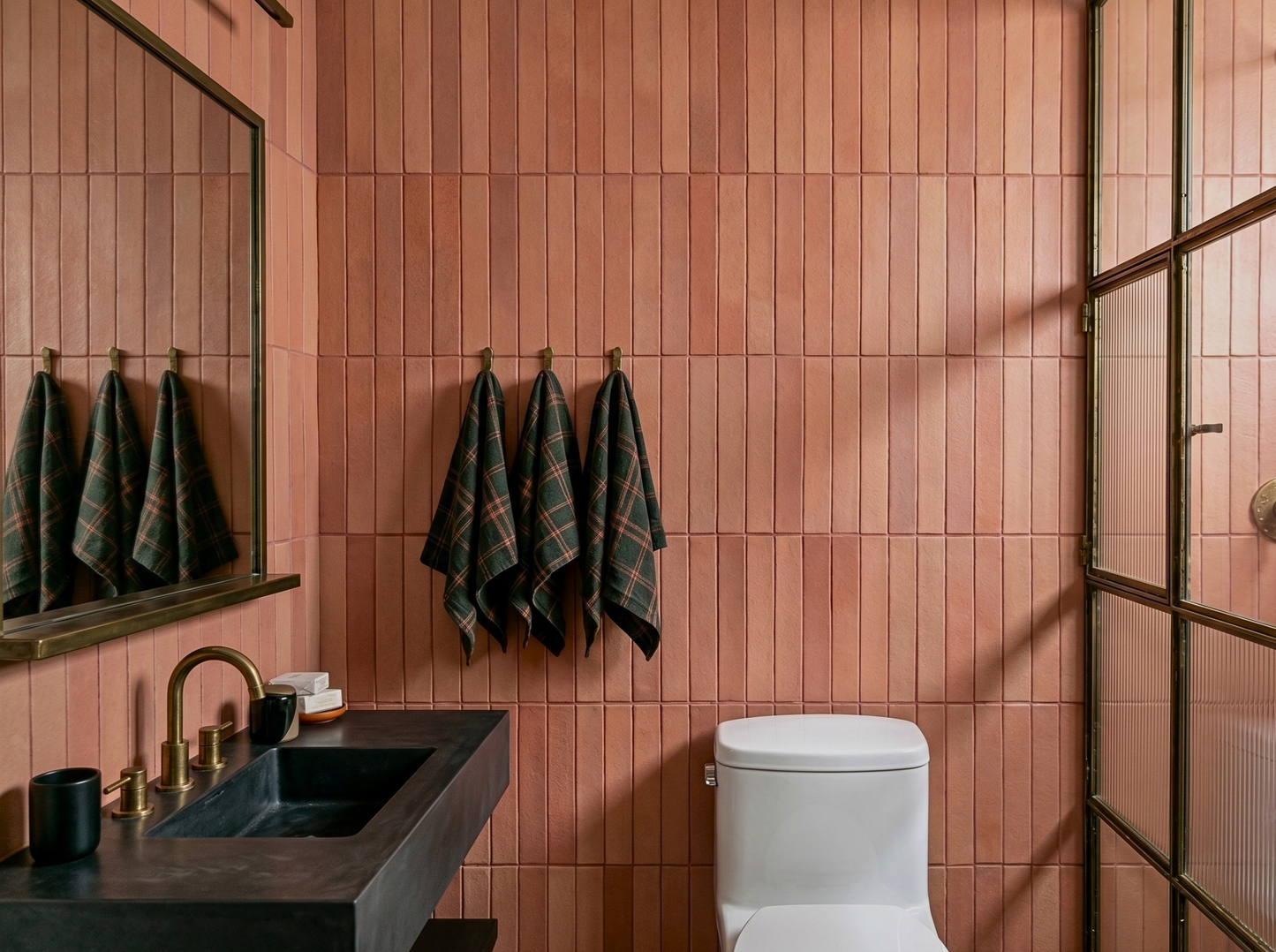 A modern bathroom interior with pinkish terracotta wall tiles, a black countertop and sink, a white toilet, and dark plaid towels hanging on hooks.