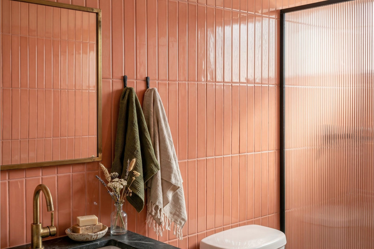 A modern bathroom interior with glossy terracotta-colored vertical wall tiles, a brass-framed mirror, a black countertop with a gold faucet, hanging towels, and a frosted glass shower door.