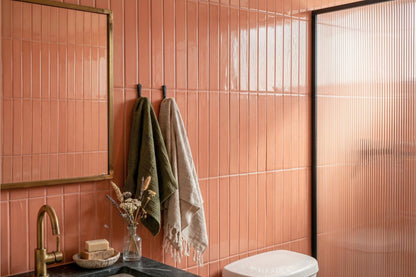 A modern bathroom interior with glossy terracotta-colored vertical wall tiles, a brass-framed mirror, a black countertop with a gold faucet, hanging towels, and a frosted glass shower door.