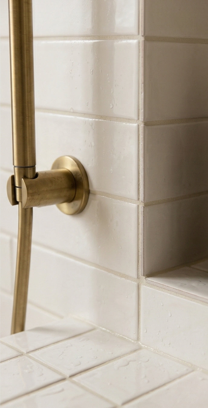 Off-white Fossil Gloss 2x5 Solid Tile by Clay Imports, wet with water droplets and a brass shower fixture in a corner.