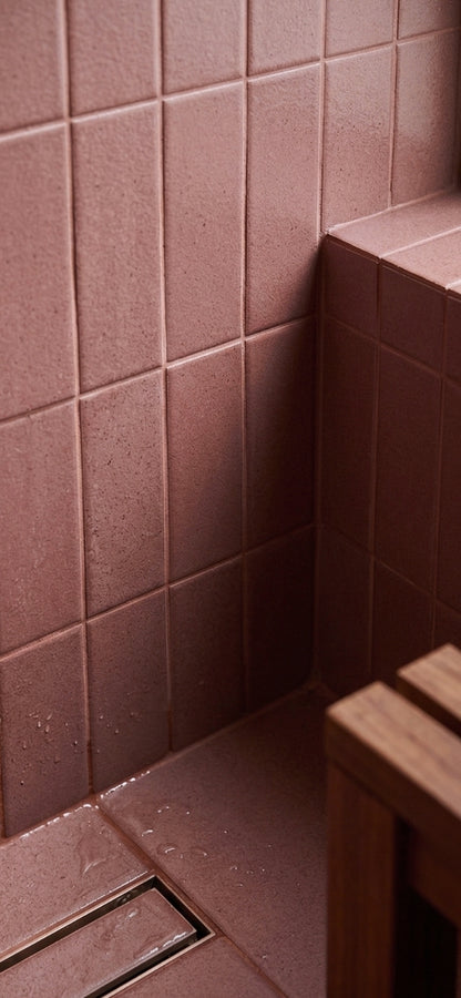 Lava Rock Matte 2x5 solid tile by Clay Imports, dusky rose with speckled texture in a shower corner with a linear drain and wood bench.