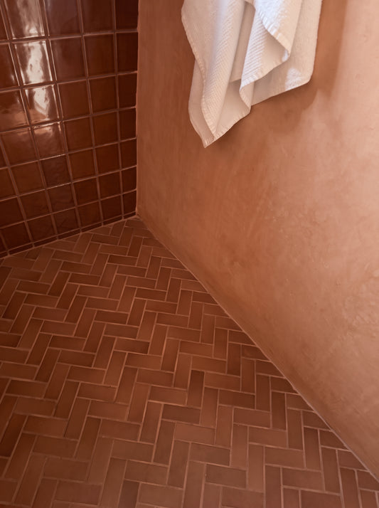 Terracotta brown herringbone tile floor and matching wall tiles in a rustic bathroom setting, with a white towel hanging on the wall.