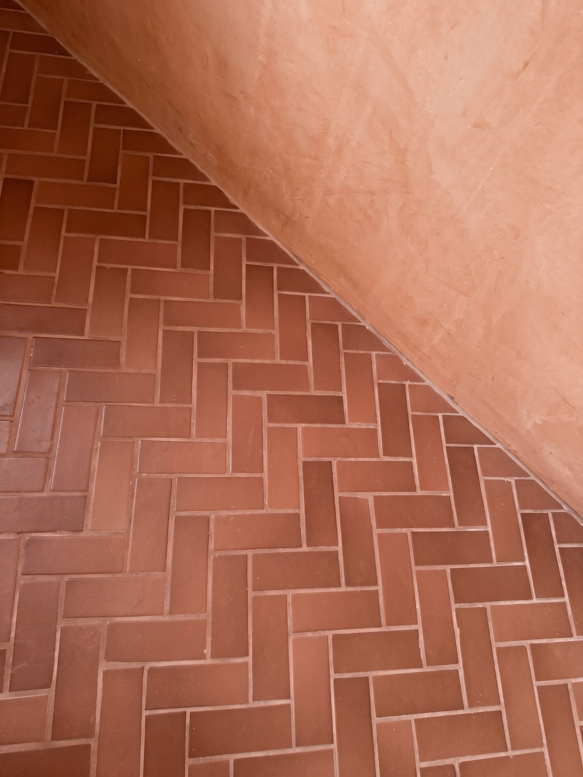 Red terracotta tiles laid in a classic herringbone pattern meet a warm peach-toned plastered wall.