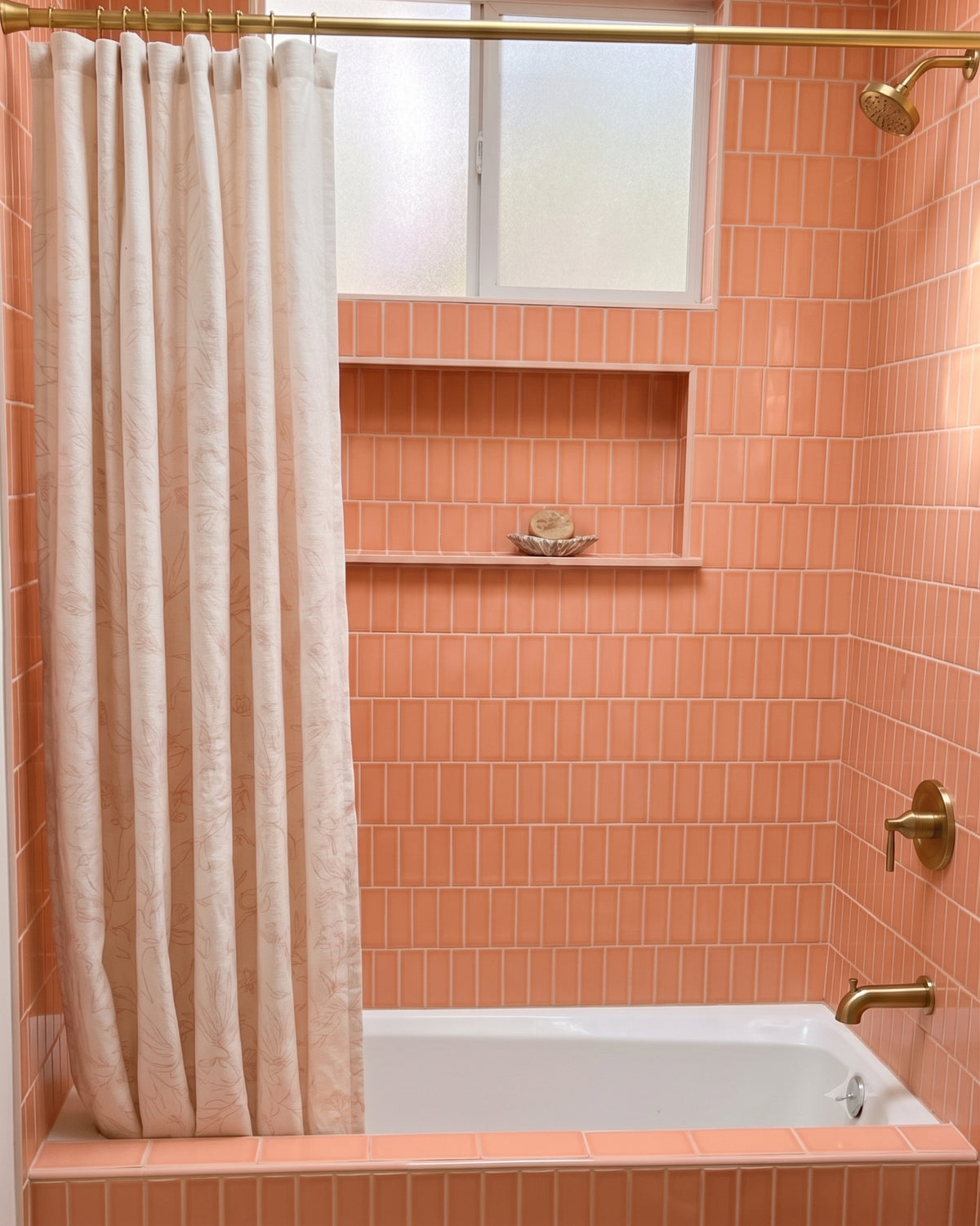 A bathroom scene with peach-orange subway tile walls, a white bathtub, a cream shower curtain, and brass fixtures. A built-in shelf holds two dark bottles and a rolled towel.