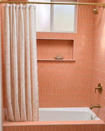A bathroom scene with peach-orange subway tile walls, a white bathtub, a cream shower curtain, and brass fixtures. A built-in shelf holds two dark bottles and a rolled towel.