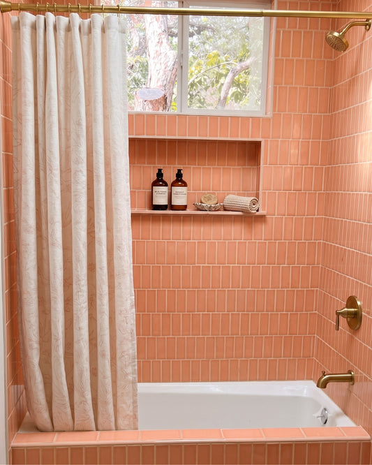A bathroom scene with peach-orange subway tile walls, a white bathtub, a cream shower curtain, and brass fixtures. A built-in shelf holds two dark bottles and a rolled towel.