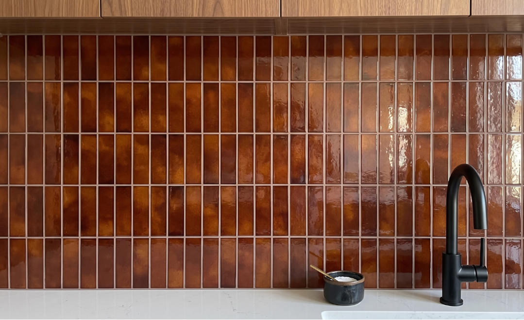 A glossy brown glass tile backsplash in a kitchen setting, paired with a matte black faucet and a light stone countertop.
