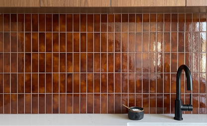 A glossy brown glass tile backsplash in a kitchen setting, paired with a matte black faucet and a light stone countertop.