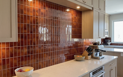 A glossy, reflective brown tile backsplash installed on a kitchen wall, paired with light beige wood cabinets and a white countertop with kitchen appliances.