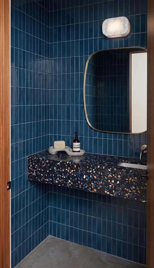 A modern bathroom vanity featuring deep blue glossy wall tiles, a floating dark speckled terrazzo countertop, a large rounded rectangular mirror with a gold frame, and a light fixture above.