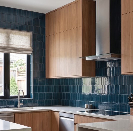 Modern kitchen interior with light wood cabinets, glossy dark blue subway tile backsplash, white countertops, a built-in sink, cooktop, and stainless steel range hood.