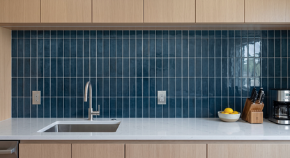 A modern kitchen backsplash featuring glossy dark blue rectangular tiles in a grid pattern, installed behind a white countertop with a stainless steel sink and faucet.